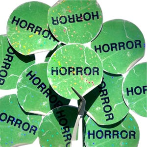 May include: A pile of green round stickers with the word "HORROR" printed in black letters. The stickers have a glittery finish.