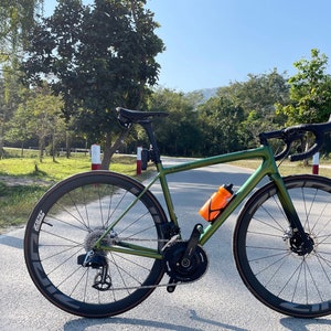 May include: A green and black road bike with a black saddle, black handlebars, and black wheels. The bike is parked on a paved road with a green grassy area and trees in the background.