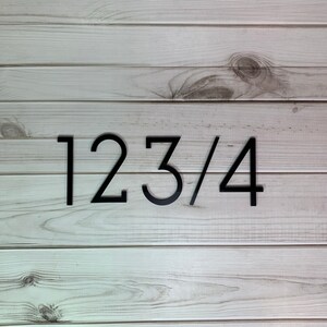 May include: Black house numbers 123/4 against a light wood background. The numbers are modern and sleek, with a minimalist design. The wood has a natural grain and a light, neutral color.