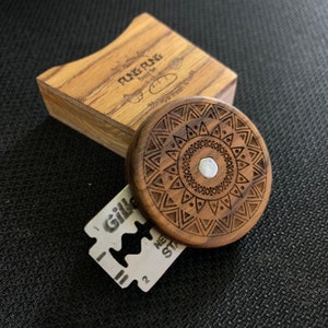 May include: A wooden round disc with a geometric pattern engraved on the surface. The disc has a silver metal center piece. The disc is sitting on a dark surface next to a razor blade.