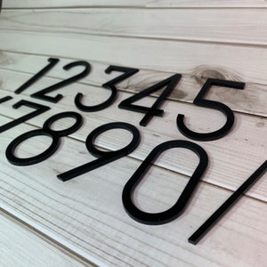 May include: A collection of black, modern house numbers, including the digits 1 through 9 and a slash. The numbers are displayed against a light wood-grain background. The numbers are in a sans-serif font.
