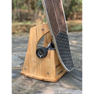 May include: A wooden skateboard stand with a skateboard resting on it. The stand is made of light brown wood and has a curved base. The skateboard is black and has a grip tape surface. The stand has the word "MANUELA" printed on it.