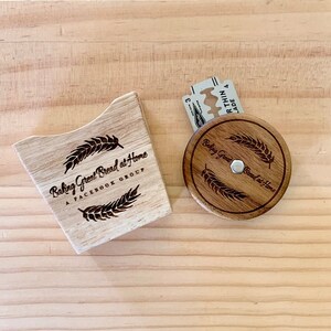 May include: A wooden box with the text "Baking Great Bread at Home A FACEBOOK GROUP" and a wheat design. A wooden round lid with the text "Baking Great Bread at Home" and a wheat design. A silver razor blade is on top of the lid.