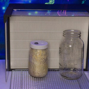 May include: Two glass jars with lids, one filled with a beige substance, sitting on a wire rack in front of a white filter inside a black frame.