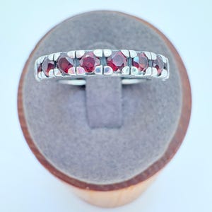 May include: A silver ring with a row of seven round, deep red gemstones. The ring is set in a silver band with a textured design. The ring is displayed on a gray velvet ring holder.