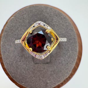 May include: A gold and silver ring with a large, square-cut, deep red gemstone at the center. The gemstone is surrounded by a gold frame with small, clear and pink accent stones. The ring is set on a gray surface.
