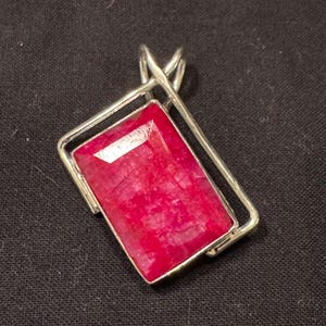 May include: A rectangular pendant featuring a vibrant, faceted red gemstone set within a polished silver-toned metal frame. The frame has a geometric design with a triangular bail at the top for attachment to a chain or cord.