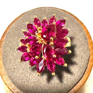 May include: A gold ring featuring a cluster of numerous marquise-cut and pear-cut deep pinkish-red gemstones arranged in a floral pattern. The stones are set in a dense, multi-layered formation that radiates outward from the center of the ring.