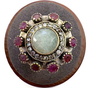 May include: A silver-toned ring with a large, pale green center stone. The ring is encircled by a row of small, clear stones and surrounded by alternating ruby-colored stones. The ring is set on a gray surface.