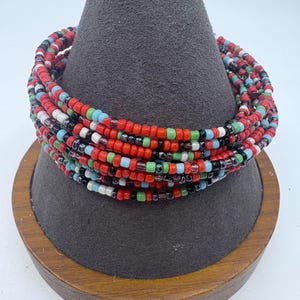 May include: A multi-strand beaded bracelet in a variety of colors, including red, black, white, green, and blue. The bracelet is displayed on a gray, cone-shaped stand with a wooden base. The beads are small and round.