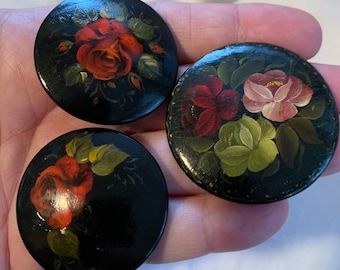 May include: Three round black wooden buttons featuring hand-painted floral designs. Two buttons display a single red rose with green leaves, while the larger button features a cluster of red, pink, and light green flowers with foliage.