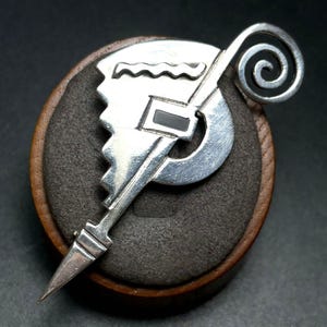May include: A silver brooch featuring a geometric, stepped design with a spiral top, a wavy line detail, and a central rectangular black inlay. The metal surface is polished and reflective, showcasing a sharp, pointed bottom edge and a curved frame.