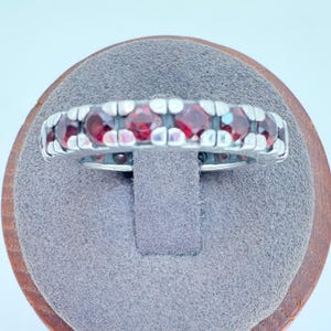 May include: A silver ring with a row of vibrant red gemstones. The gemstones are set in a channel setting, creating a continuous band of color. The ring is displayed on a gray surface, showcasing its design and craftsmanship.