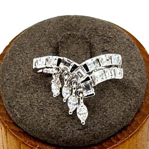 May include: A silver ring with a unique design. The ring features a double band with square and rectangular clear stones. Dangling from the ring are several teardrop-shaped clear stones. The ring is displayed on a brown surface.