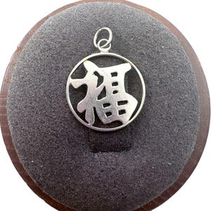 May include: A circular silver pendant featuring a cutout of the Chinese character for good fortune, known as Fu. The pendant has a polished metallic finish and includes a small silver jump ring at the top for attachment to a chain or cord.