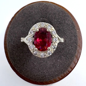 May include: A silver ring with a large, oval-cut, deep red gemstone surrounded by a halo of smaller, clear stones. The ring is set on a dark gray, circular display stand with a wooden edge.