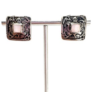 May include: A pair of square-shaped silver earrings featuring an oxidized finish with intricate, raised scrollwork patterns and a central, smooth, pale pink square inlay. The earrings are displayed on a horizontal silver-toned metal bar.