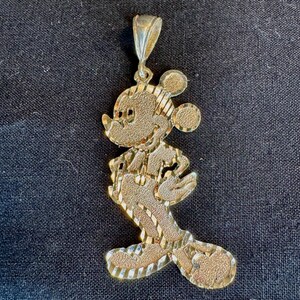 May include: A gold-toned metal pendant shaped like a cartoon mouse character in profile. The surface features a textured, diamond-cut finish with alternating polished diagonal stripes, and it includes a tapered bail at the top for hanging.
