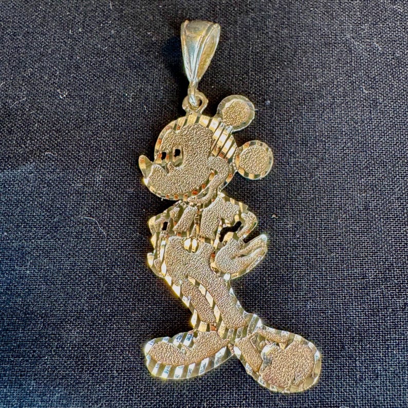May include: A gold-toned metal pendant shaped like a cartoon mouse character in profile. The surface features a textured, diamond-cut finish with alternating polished diagonal stripes, and it includes a tapered bail at the top for hanging.