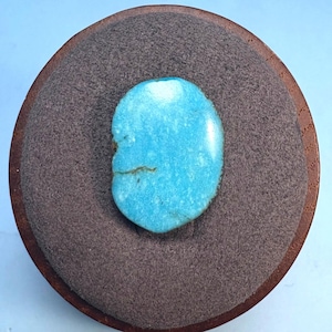 May include: A smooth, oval-shaped turquoise stone with a light blue color. The stone is set on a brown, textured surface.