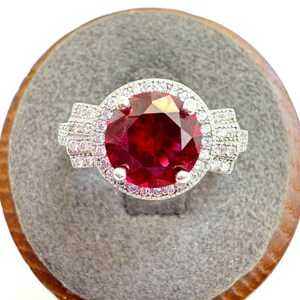 May include: A silver ring featuring a large, round, faceted, deep red gemstone, surrounded by a halo of smaller, clear stones. The ring's band has additional clear stones on each side, set on a gray surface.