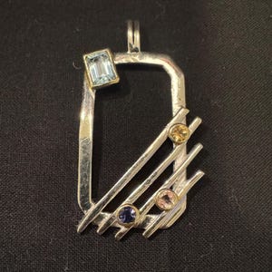 May include: A rectangular silver pendant featuring a top-left emerald-cut light blue gemstone set in gold. Three diagonal silver bars cross the lower right section, each accented with a small round gemstone in yellow, pink, and dark blue.