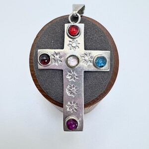 May include: A silver cross pendant featuring five circular cabochon stones in red, blue, purple, and clear colors. The metal surface is decorated with stamped sunburst patterns along the vertical and horizontal arms of the cross shape.