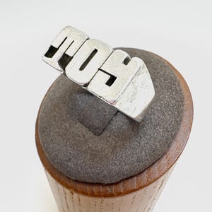 May include: A silver ring with the letters "MOS" in a bold, blocky font. The ring is displayed on a gray, cylindrical stand with a wooden base. The ring's design is simple and modern, with a slightly textured surface.