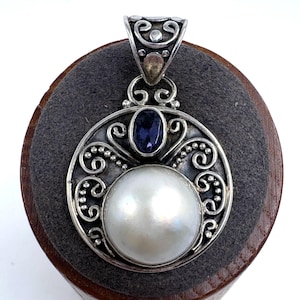 May include: A circular silver pendant featuring a large, round white pearl at the center, topped with an oval dark blue gemstone. The silver frame is decorated with intricate scrollwork and bead detailing, finished with a decorative silver bail.