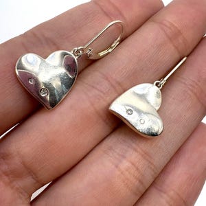 May include: A pair of silver heart-shaped earrings. Each earring features a heart design with a small, round, clear gemstone. The earrings have a lever-back closure. The earrings are displayed against a light background.