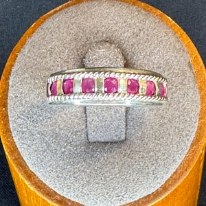 May include: A silver band ring featuring a row of seven round, vibrant pinkish-red rubies set between two twisted rope-style silver borders. The metal has a polished finish, and the stones are evenly spaced along the center of the band.