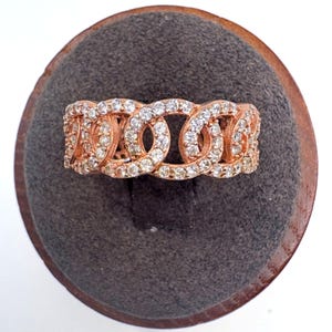 May include: A rose gold band ring featuring a continuous chain-link design. The surface of the metal links is encrusted with numerous small, clear, sparkling stones that cover the entire visible front portion of the ring's band.