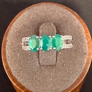 May include: A silver ring featuring three oval-cut green gemstones arranged horizontally in the center. The band is split into two thin, textured arms on each side, with small clear stones embedded along the metal surface of the band.