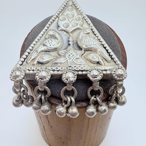 May include: A triangular silver-toned metal pendant featuring an embossed design of two birds facing a central floral motif. The base of the triangle is adorned with five small, dangling spherical charms attached by metal rings.