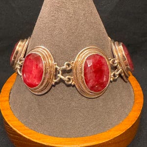 May include: A link bracelet featuring four oval-shaped, faceted red stones set in silver-toned metal frames. Each stone is surrounded by a decorative rope-like border, and the individual settings are connected by small silver-toned metal jump rings.