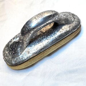 May include: An antique silver-plated clothes brush with an ornate floral design. The brush features a curved handle with engraved initials and a rectangular base with a tan-colored pad. The brush is likely from the Victorian era.