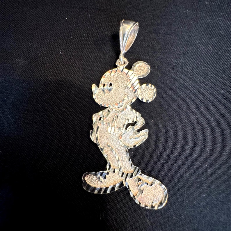 May include: A silver-toned metal pendant shaped like a cartoon mouse character. The surface features a combination of textured, matte, and polished finishes with diagonal striped detailing throughout the body and ears, including a top bail loop.