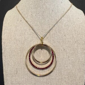May include: A gold-toned necklace with three interlocking circles. The two outer circles are plain, while the middle circle is adorned with small, red gemstones. The necklace hangs from a delicate chain.