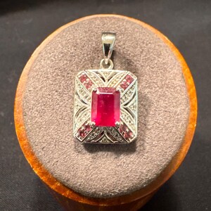 May include: A rectangular silver pendant featuring a large, central emerald-cut red gemstone. The frame is decorated with smaller red stones arranged in an X-pattern, accented by tiny clear stones along the silver metalwork and a top bail.