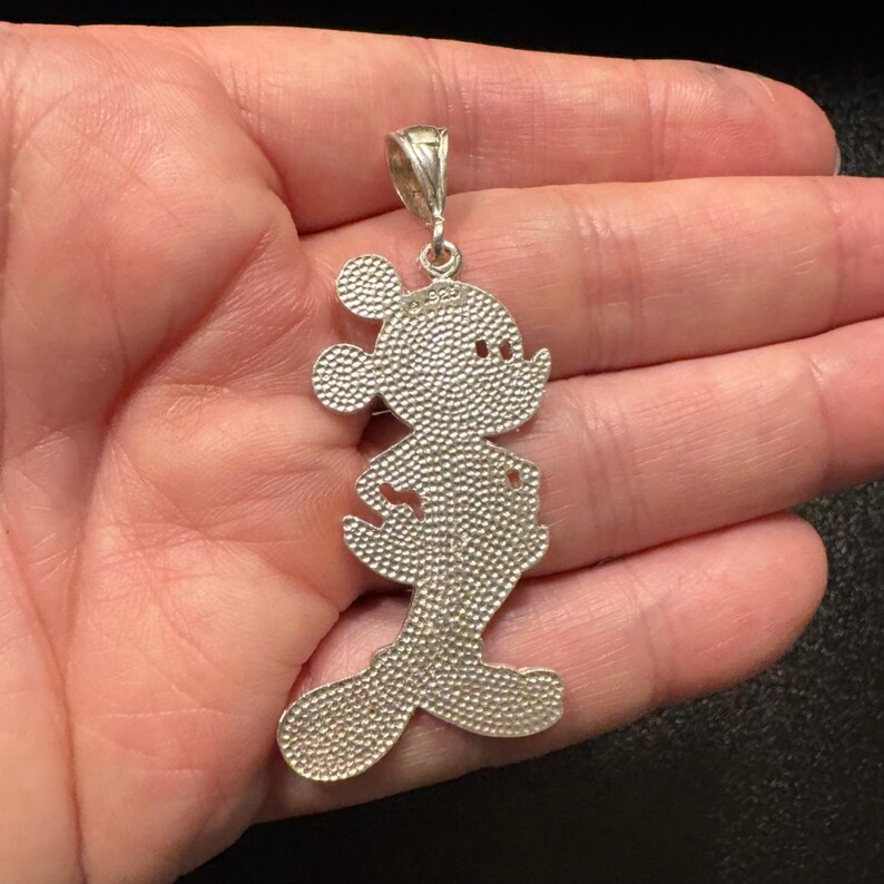 May include: A silver-toned metal pendant shaped like the silhouette of a cartoon mouse character. The surface features a textured, stippled finish throughout the body and ears. A small metal bail is attached to the top for hanging on a chain.