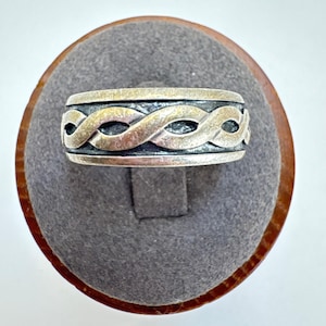 May include: A silver-toned ring with an interwoven design. The ring features a band with a repeating pattern of intertwined loops, creating a textured appearance. The ring is displayed on a gray, oval-shaped surface.