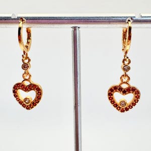 May include: Gold-toned heart-shaped earrings with red and clear gemstones. The earrings have a dangling design with a small gemstone at the top and a heart-shaped pendant. The heart is outlined with red gemstones and has a clear gemstone in the center.
