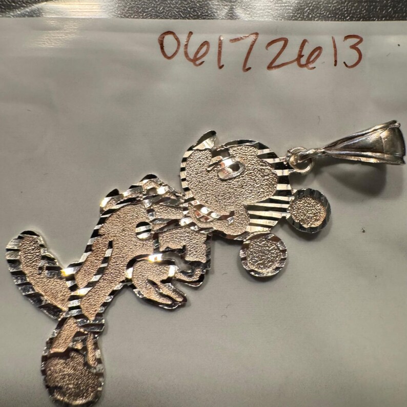May include: A two-tone silver and rose-coloured metal pendant featuring a textured, stylised cartoon character design with a bail at the top. The piece has a diamond-cut finish with alternating shiny and matte surfaces. The number 06172613 is visible.