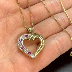 May include: A gold heart-shaped pendant necklace featuring a series of small, round, vibrant red rubies and tiny clear accent stones set along one side of the heart frame. The pendant hangs from a delicate, thin gold box chain necklace.