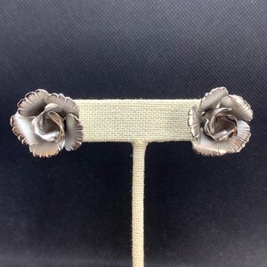 May include: A pair of silver-toned rose-shaped earrings. The earrings are displayed on a beige stand against a dark background. The rose petals have a textured appearance, and the overall design is detailed and elegant.