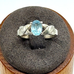 May include: A silver ring featuring an oval, light blue gemstone. The ring's band has a braided design, with the gemstone set in the center. The ring is displayed on a dark gray, textured surface, set on a wooden base.