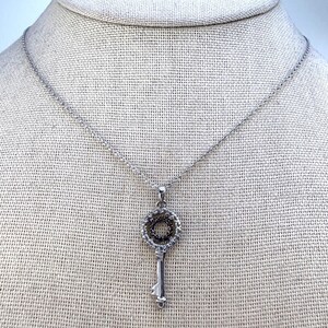 May include: A silver-toned necklace featuring a key-shaped pendant. The pendant has a circular top encrusted with small clear stones surrounding a smaller inner circle of dark stones. The key shaft is smooth silver metal with a simple notched end.