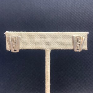 May include: Silver-toned geometric stud earrings. Each earring features a stylized, open design with a rectangular shape and a Greek key pattern. The earrings are displayed on a beige fabric-covered stand against a dark blue background.