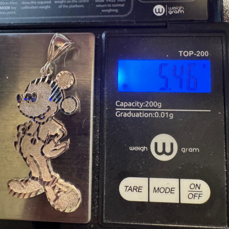 May include: A silver-toned metal pendant shaped like a cartoon mouse character with a textured finish. The pendant is placed on a digital scale that displays a weight of 5.46 grams. The scale has a black frame and a blue backlit digital display.