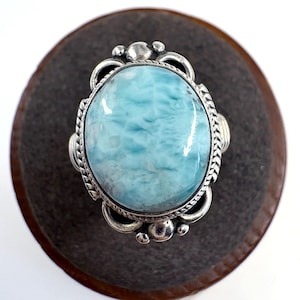 May include: A silver ring featuring a large, oval-shaped, light blue Larimar gemstone with a marbled texture. The setting is crafted from polished silver with a twisted rope border and decorative metal loops and spheres around the stone.